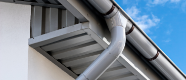 Gray rain gutter system with downspout installed on white house exterior roof